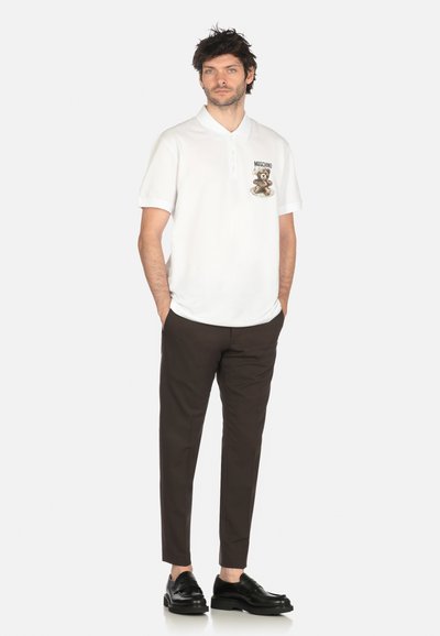 Uomo in piedi con le mani in tasca, indossa una polo bianca con logo dell'orso, pantaloni marrone scuro e mocassini neri su sfondo bianco.