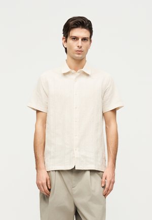 Jeune homme portant une chemise texturée blanche cassée à manches courtes et boutonnée ainsi qu'un pantalon beige, debout devant un fond clair uni.