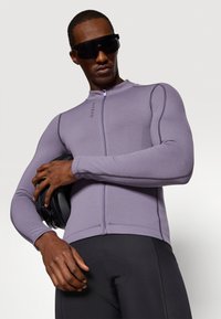 Jersey de ciclismo de manga larga en tela púrpura clara, con cremallera frontal y costuras planas, combinado con pantalones cortos de ciclismo negros y sosteniendo un casco.