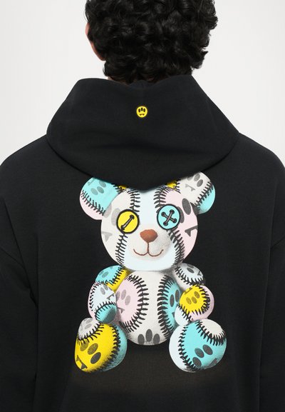 Persona che indossa una felpa nera con cappuccio decorata con un grafico di un orsetto colorato ricamato e un piccolo icono di impronta gialla sul retro del cappuccio.