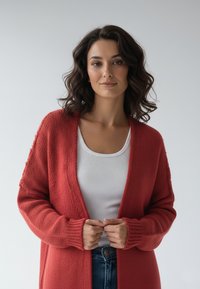 Czerwony dziergany sweter zapinany na guziki z długimi rękawami, otwarty z przodu, wykonany z teksturowanej tkaniny, noszony na białym prążkowanym topie na ramiączkach, w połączeniu z niebieskimi dżinsami.