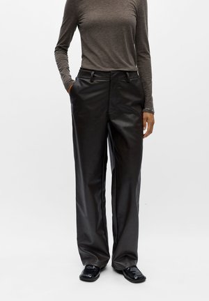Personne portant un pantalon en cuir noir à jambes larges, un haut marron moulant à manches longues et des chaussures noires à bout carré, debout les mains dans les poches.