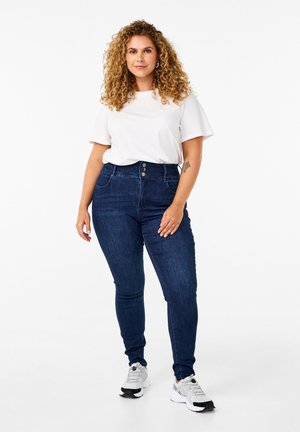 Jeans de tiro alto azul oscuro con cierre de tres botones, dos bolsillos frontales y corte ajustado, combinados con una camiseta blanca y zapatillas.