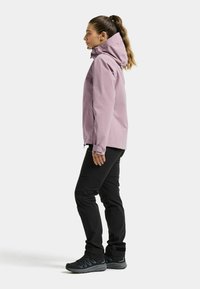 Woman standing sideways wearing a light purple hooded jacket, black pants, and black hiking shoes against a plain white background.