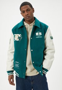 Varsityjack in teal wolblend met witte lederen mouwen. Voorzien van geborduurde Tokyo graphics, drukknoopsluiting en geribde manchetten.