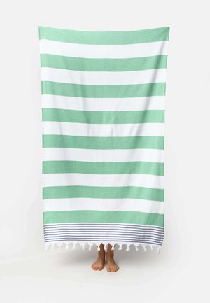 Grün-weiß gestreiftes Strandtuch mit Fransen, gehalten von einer Person, die nur ihre barfüßigen Füße unter dem Handtuch zeigt.
