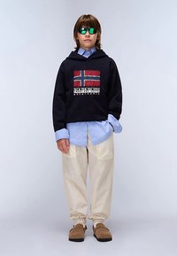 Sudadera negra con diseño de la bandera de Noruega, combinada con una camisa azul claro abotonada, pantalones amplios color crema y zapatos marrones sin cordones. Gafas de sol verdes.