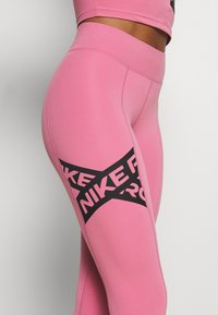 Leggings rosas hechos de un tejido elástico con una cinturilla alta y un gran logo negro de "NIKE" en el costado, con una textura suave.