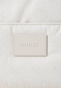 Tissu blanc texturé avec un motif subtil. Présente une plaque de logo rectangulaire en relief qui indique "GUESS" en couleur gris clair.