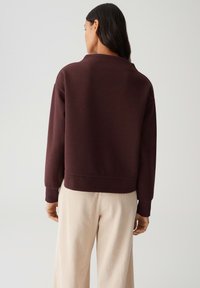 Vrouw met lang donker haar, gekleed in een donkerbruin sweatshirt en lichtbeige geribbelde wijde broek, met de rug naar de camera gekeerd tegen een effen achtergrond.