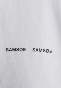 White cotton shirt featuring two black printed instances of "SAMSØE" in a bold, sans-serif font, positioned horizontally on the front.