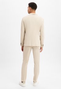 Helles beiges Anzugensemble mit einem strukturierten Blazer mit einer Schlitze und passenden Hosen. Glattes Material und taillierte Passform. Schlichte weiße Sneakers.