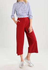 Light blue striped shirt with pearl accents, red wide-leg trousers, and silver slip-on shoes. Fabrics are smooth and lightweight.