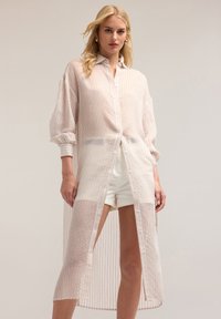 Camicia leggera, lunga e bianca con righe rosse verticali, chiusura con bottoni e maniche a sbuffo, caratterizzata da una trama trasparente e fessure laterali.