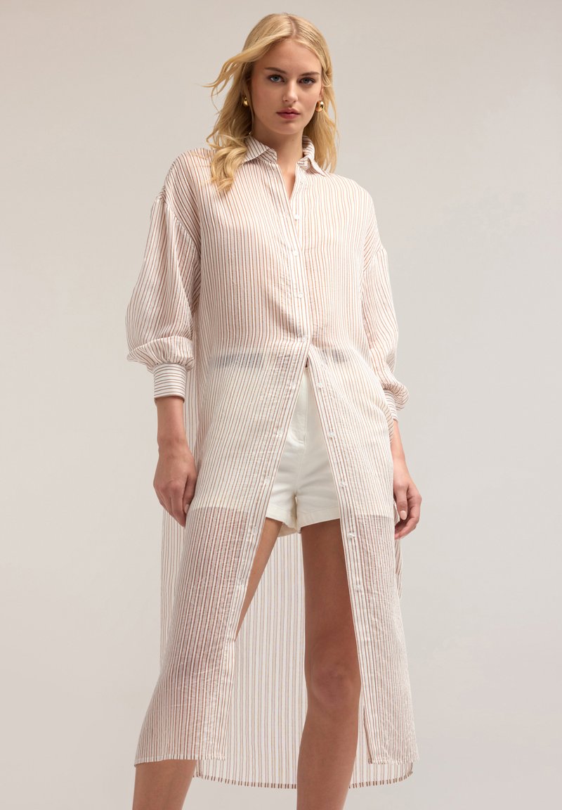 Camicia leggera, lunga e bianca con righe rosse verticali, chiusura con bottoni e maniche a sbuffo, caratterizzata da una trama trasparente e fessure laterali.