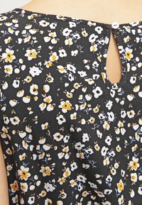 Blouse noire à fleurs avec de petites fleurs blanches et jaunes, détail en forme de trou de serrure au dos avec une fermeture à bouton, tissu léger.