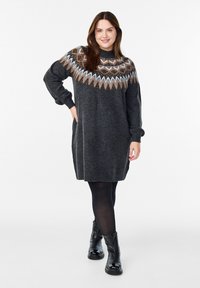 Robe tricotée gris foncé avec un empiècement à motifs en orange, blanc et marron. Dotée de manches longues et d'une coupe décontractée, elle est portée sur des collants noirs et des bottes.