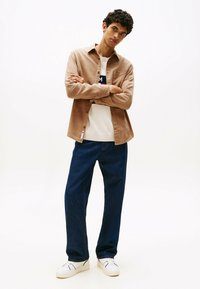 Light brown button-up shirt over a white sweater, paired with dark blue jeans and white sneakers. Model posing with arms crossed.