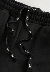 Mustad mustadid sisaldavad elastset vööd, millel on mustast kangast kinnitusnöör, millel on valgete tähtedega märge "self." Sujuv tekstuur, kodune disain.