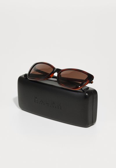Brązowe okulary przeciwsłoneczne z przyciemnianymi szkłami spoczywające na zamkniętym czarnym etui Ralph Lauren na jednolitym jasnym tle.