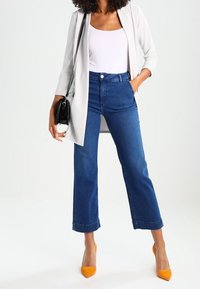 Jean bleu taille haute à jambes larges, assorti d'un débardeur blanc et d'un blazer gris clair, rehaussé par des talons orange et un sac noir.
