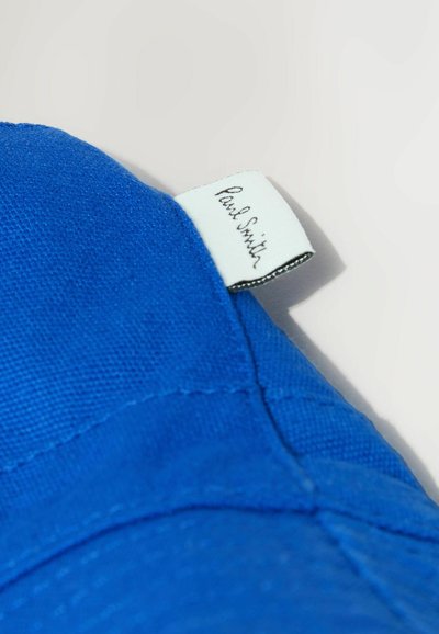 Tissu bleu avec une étiquette blanche portant « Paul Smith » en broderie cursive noire, cousue dans la couture.