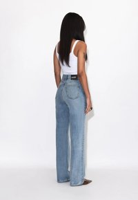 Woman with long dark hair stands facing away, wearing a white tank top and high-waisted light blue wide-leg jeans against a white background.