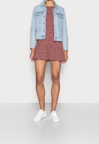 Frau in einem roten, blumigen Mini-Kleid, einer hellblauen Jeansjacke und weißen Sneakers, die vor einem schlichten weißen Hintergrund steht.