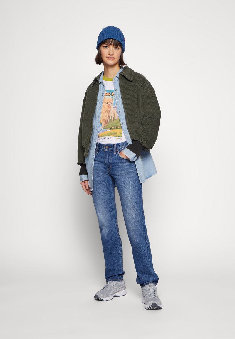 Joven con gorro azul, chaqueta de pana verde, camisa de mezclilla en capas, camiseta con gráfico de gato, jeans azules y zapatillas grises, de pie con una mano en el bolsillo.