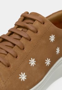 Brun mockasneaker med vit blomsterbroderi. Har runda skosnören och en texturerad yta. Vit gummisula.