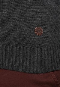 Maglione grigio scuro lavorato a maglia con orlo a coste e piccolo logo rotondo in pelle marrone etichettato "Springfield" sopra un tessuto rosso scuro.