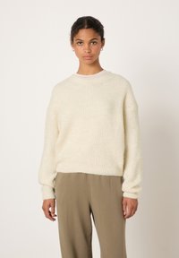 Jauna moteris stovi, dėvi kreminės spalvos megztinį, smėlio spalvos kelnes, mažus auksinius lankelius ausyse, prieš šviesų vienodą foną.