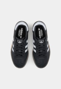 Černé tenisky z hladkého materiálu s bílými pruhy, kulatými tkaničkami a texturovanou šedou podrážkou. Logo je viditelné na jazyku boty.