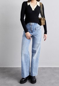 Haut noir à manches longues côtelé avec un col crème, associé à un jean large bleu clair et des bottes plateformes noires. Sac tressé marron suspendu à l'épaule.