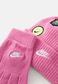 Gants en tricot roses avec texture côtelée et logo Nike blanc. Comprend un bonnet rose assorti avec un patch de visage souriant.