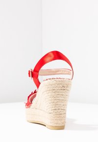 Sandalia cuña roja con una plataforma de yute tejido texturizado y una correa de cuero suave con cierre. Presenta un tacón curvado y un forro interior suave.