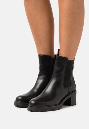 Botas de tobillo de cuero negro con un tacón grueso, paneles laterales elásticos y una suela de goma texturizada. Diseño minimalista con un detalle de logo en el lateral.