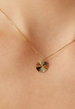 Goldkette mit einem runden Anhänger, der eine radiale Textur und eingravierte Linien aufweist, liegt auf glatter Haut. Die Kette ist zart und gliedert.