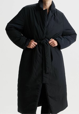 Personne attachant la ceinture d'un long manteau noir matelassé avec col haut et manches longues sur un fond uni.