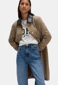 Cappotto lungo a quadri marroni con colletto in pelle nera, abbinato a una felpa grigia con testo nero e jeans blu a vita alta.