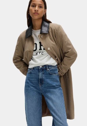 Brun ternet lang frakke med en sort læderkrave, parret med en grå sweatshirt med sort tekst og højtaljede blå jeans.
