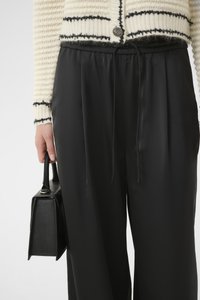 Pantaloni neri in satin con vita elastica, abbinati a un cardigan bianco testurizzato con strisce nere e un bottone rotondo. Tiene una borsa nera.