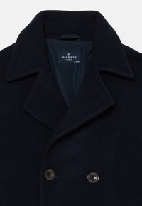 Mornarska volnena peacoat jakna z velikimi klapnicami, dvojno zapiranje s gumbi in dvema črnima gumboma z embosiranim logotipom. Notranjost je podložena.