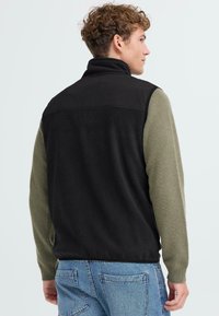 Schwarze Fleece-Weste mit grünen Strickärmeln, hohem Kragen und flachen Nähten. Verfügt über eine lockere Passform und eine weiche Textur.