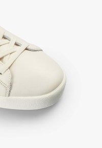 Cream-colored sneaker with a smooth leather upper, textured rubber sole, and flat cotton laces. Rounded toe and subtle stitching details.