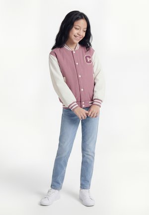 Jente som står og smiler, iført en rosa og hvit varsityjakke, lyseblå jeans og hvite joggesko mot en ensfarget hvit bakgrunn.