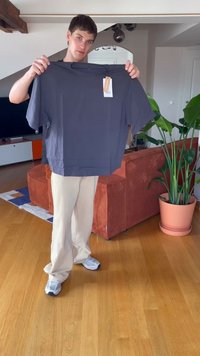 Jauns vīrietis stāv viesistabā, turēdams rokā tumši pelēku T-kreklu ar birkām, ģērbies bēšās biksēs un baltos sporta apavos.