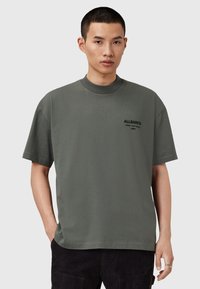 Camiseta de algodón verde oscuro con un cuello alto, mangas cortas y un estampado de logotipo en el frontal. Textura suave y diseño de corte relajado.