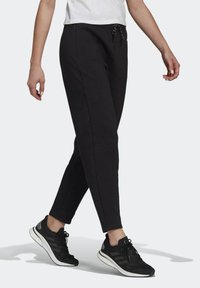 Persoon in zwarte joggingbroek en zwarte Adidas hardloopschoenen, lopend tegen een effen lichte achtergrond.