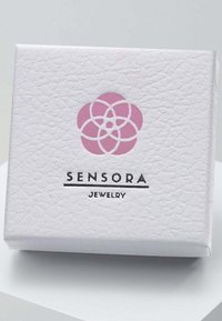 Baltā rotaslietu kaste ar teksturētu virsmu, kurai ir rozā ziedu logo un melnā krāsā izdrukāts "SENSORA JEWELRY". Kvadrātveida forma, minimālistisks dizains.